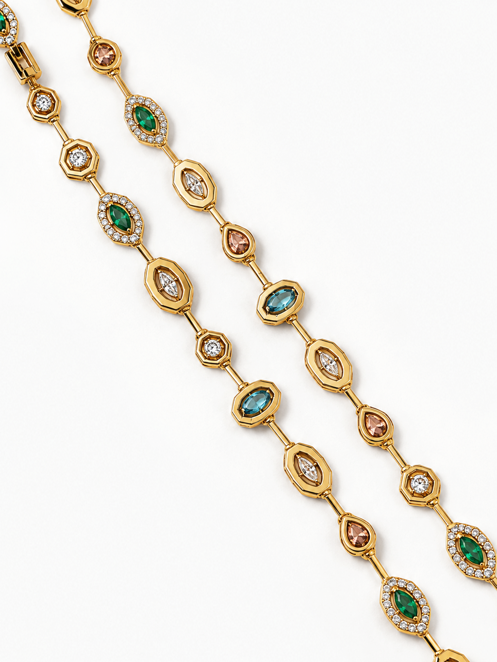 A close-up studio shot of a gold chain necklace laid diagonally on a white background. The necklace features alternating oval and marquise-shaped links, some set with green emeralds, others with light blue topazs, and several with small peach-colored stones.
